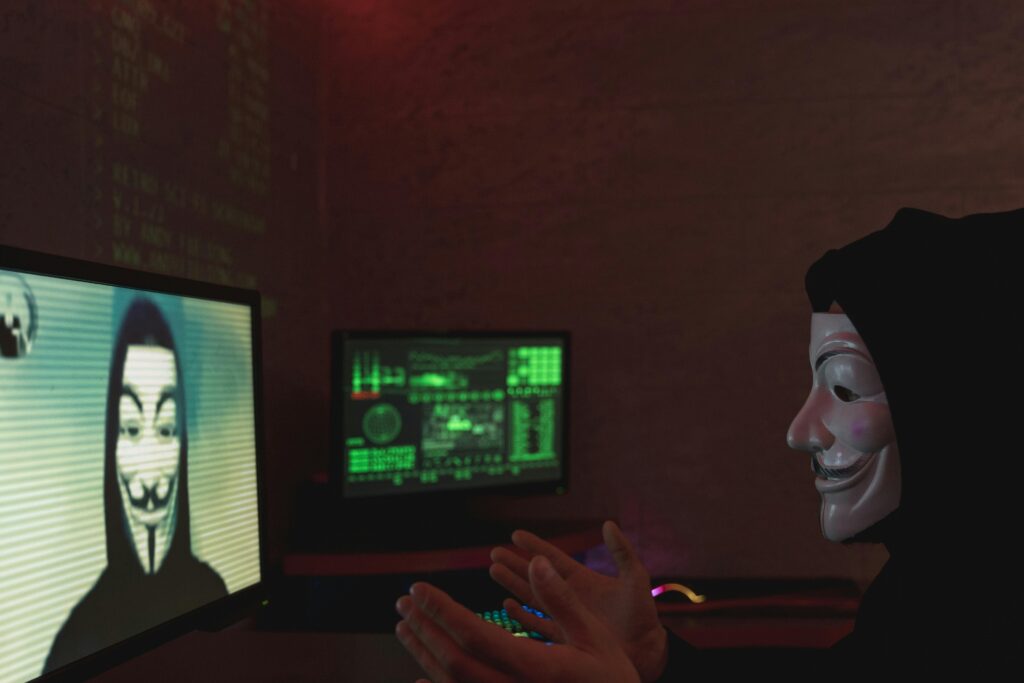 Masked individual in dark room interacting with computer screens displaying hacker imagery and code.